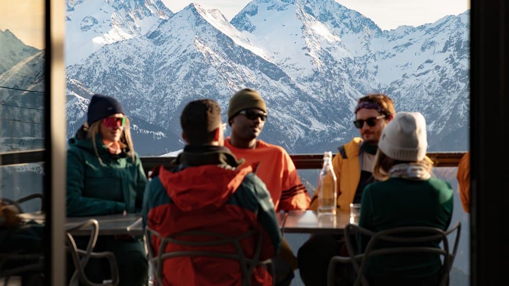 Friends at a table on a mountain terrace, high peaks behind the glass