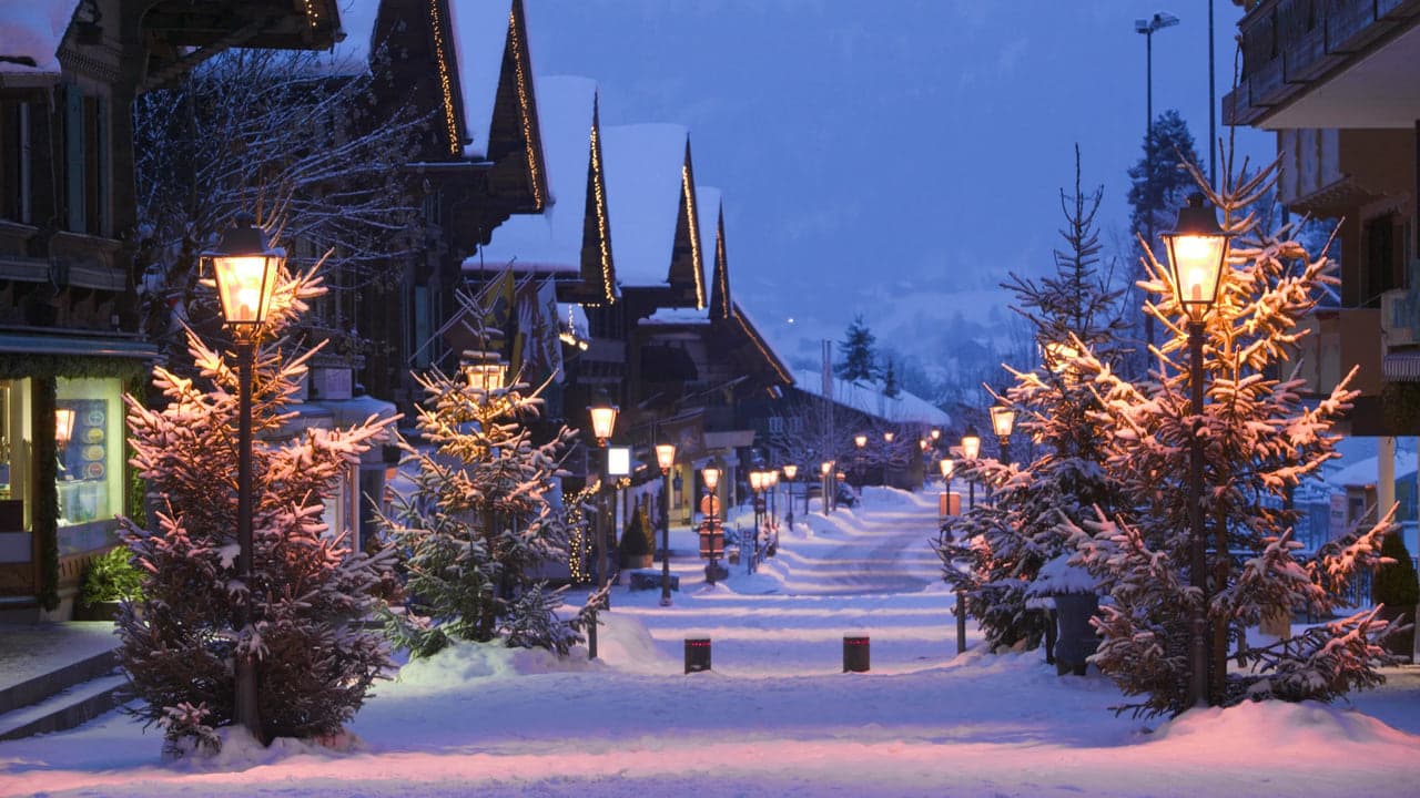 A snow covered village street at dusk lined with lit lamps and frosted trees, no people in frame