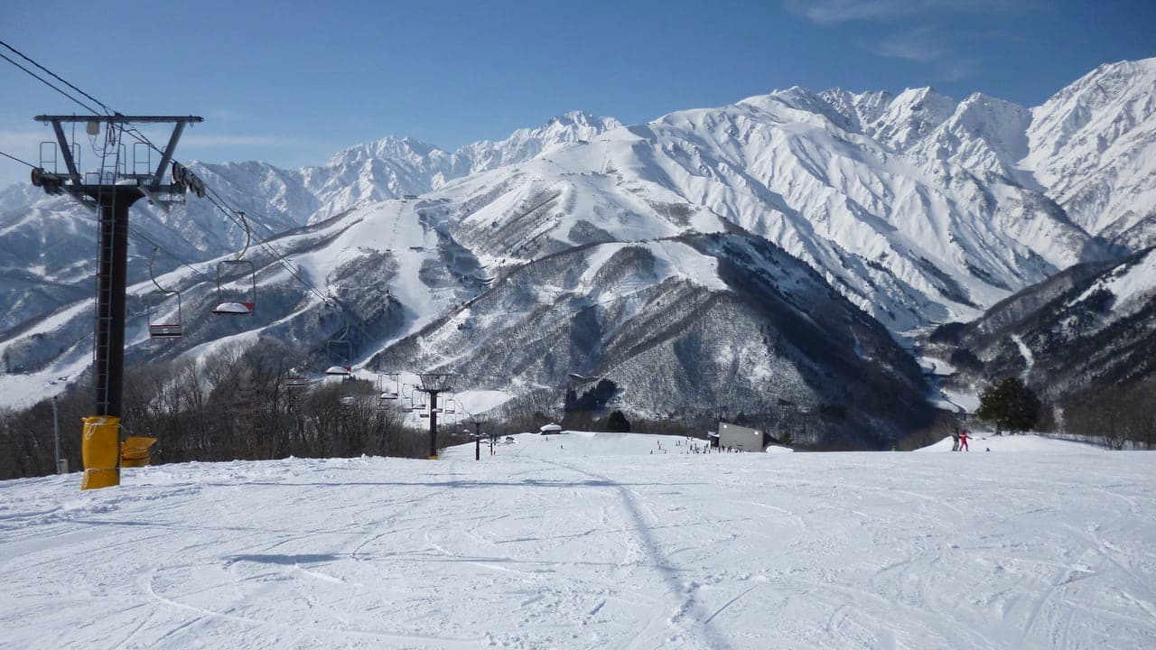 An empty groomed run below a chairlift, snowy peaks in the distance
