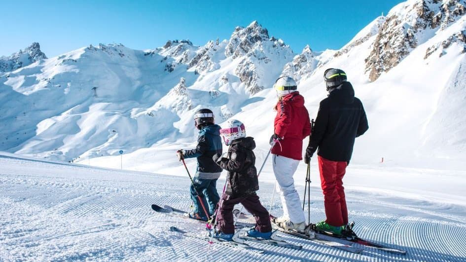 A family skiing together on a groomed run below jagged peaks