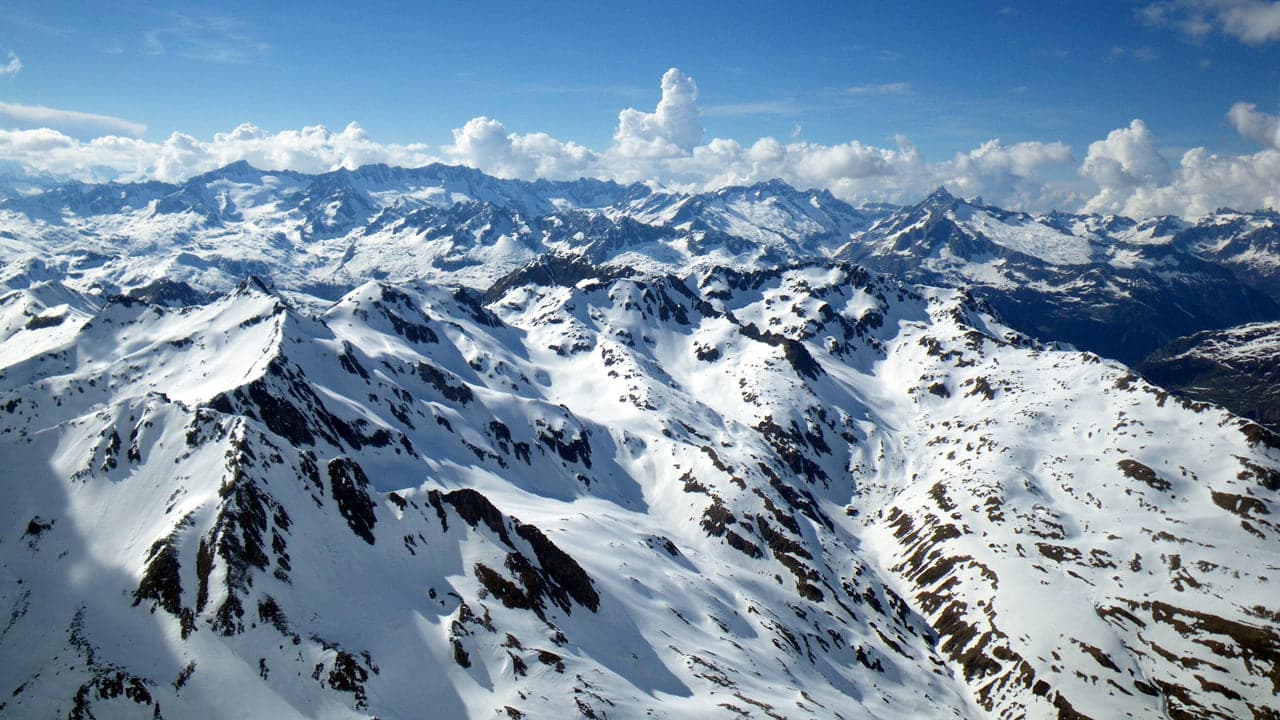 Wide aerial view of snow covered alpine peaks under a blue sky with scattered clouds